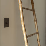 Interruptor articulado individual Bronze montado numa parede de cor creme à esquerda com uma escada de bambu à direita. 